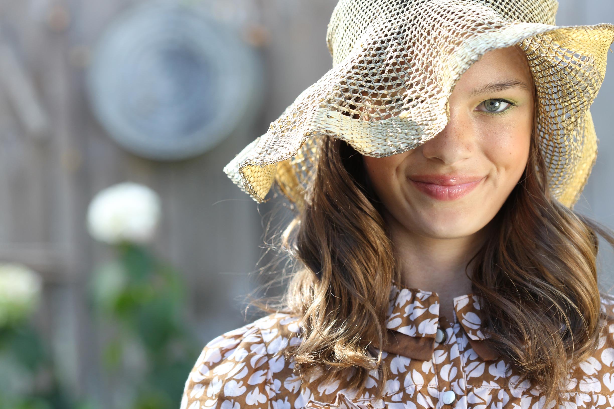 A young woman with long brown hair wearing a straw hat with a wide brim, a brown and white patterned blouse, standing outdoors with a blurred garden background.